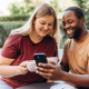 A black man holds a phone as a white woman sitting next to him looks at the screen and both smile, an example of an imaged used in positive psychology web design. Both have coffee cups in their hands.