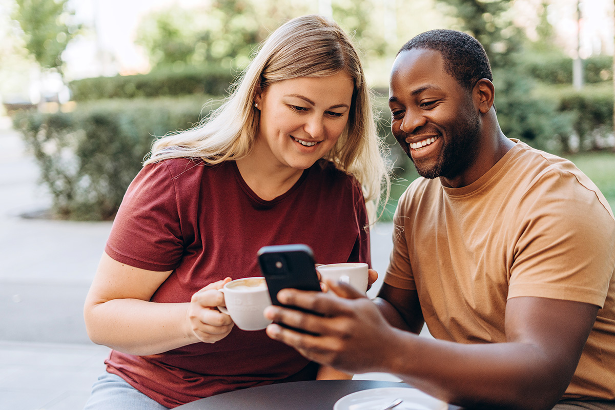 A black man holds a phone as a white woman sitting next to him looks at the screen and both smile, an example of an imaged used in positive psychology web design. Both have coffee cups in their hands.