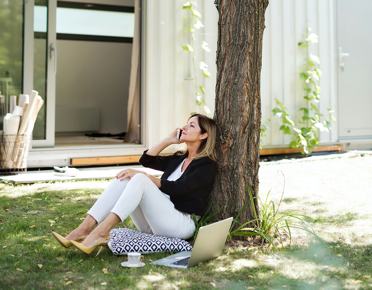 Woman sitting outside leaning against a tree with a house in the background while she talks on the phone, pondering her website's scalable design