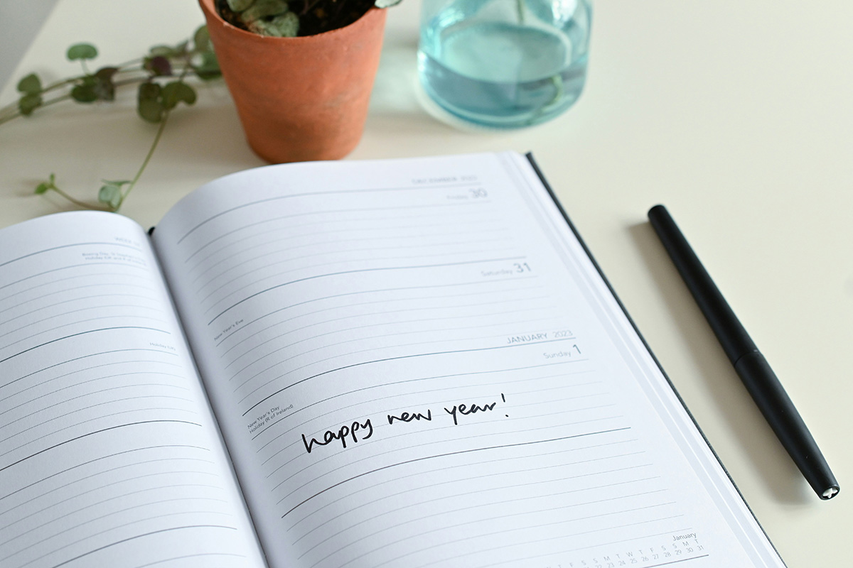 An agenda open on a desk with "Happy New Year!" written on the middle of the right page in black ink. A black pen sits next to the agenda.